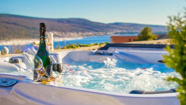 A hot tub with a bottle of champagne, a glass, and a remote control, set against a scenic backdrop of hills and a body of water. - Villa Antonia (Krk)