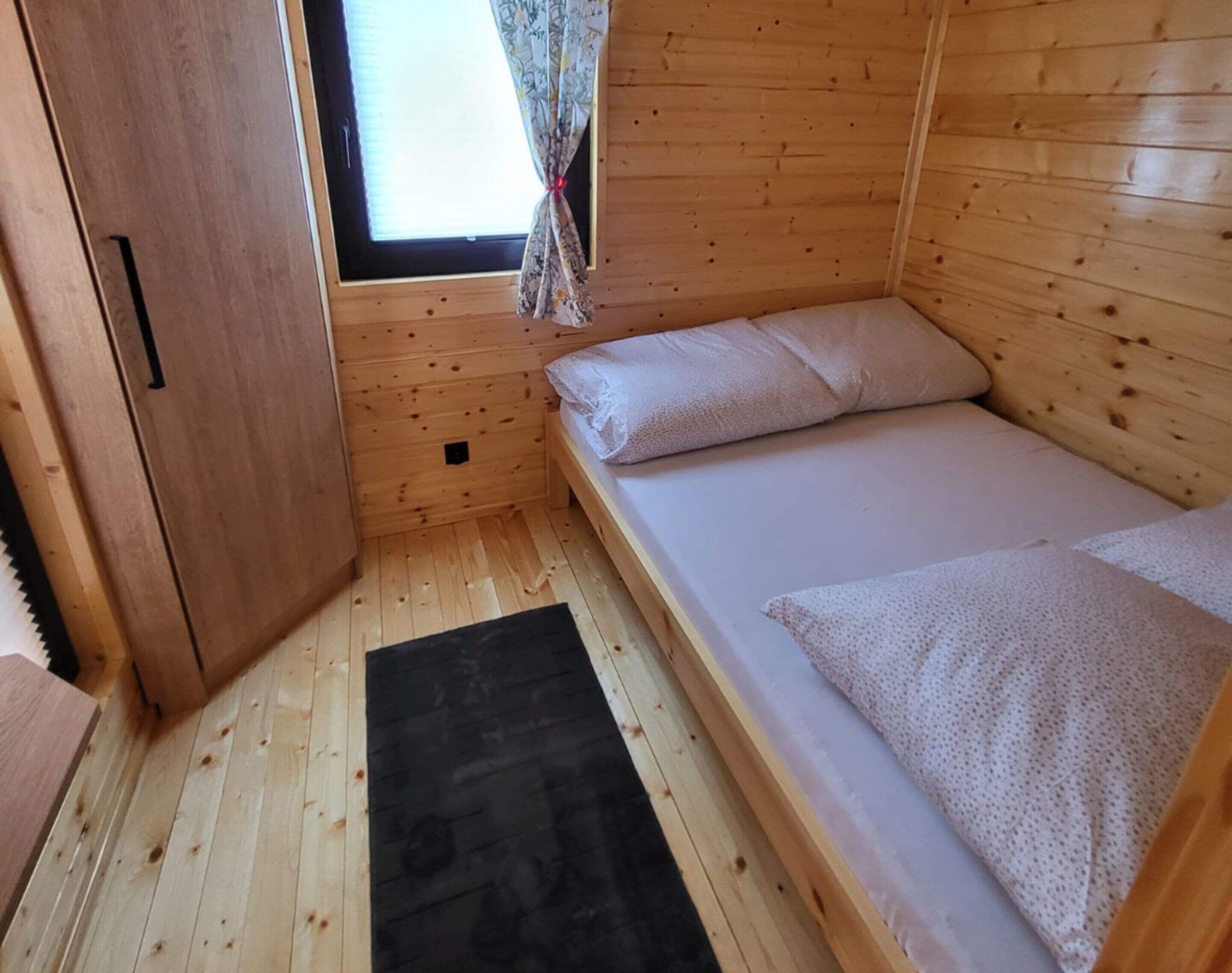 A small wooden cabin room with a single bed, a window with curtains, and a door.