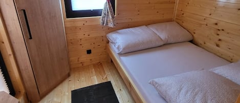 A small wooden cabin room with a single bed, a window with curtains, and a door.