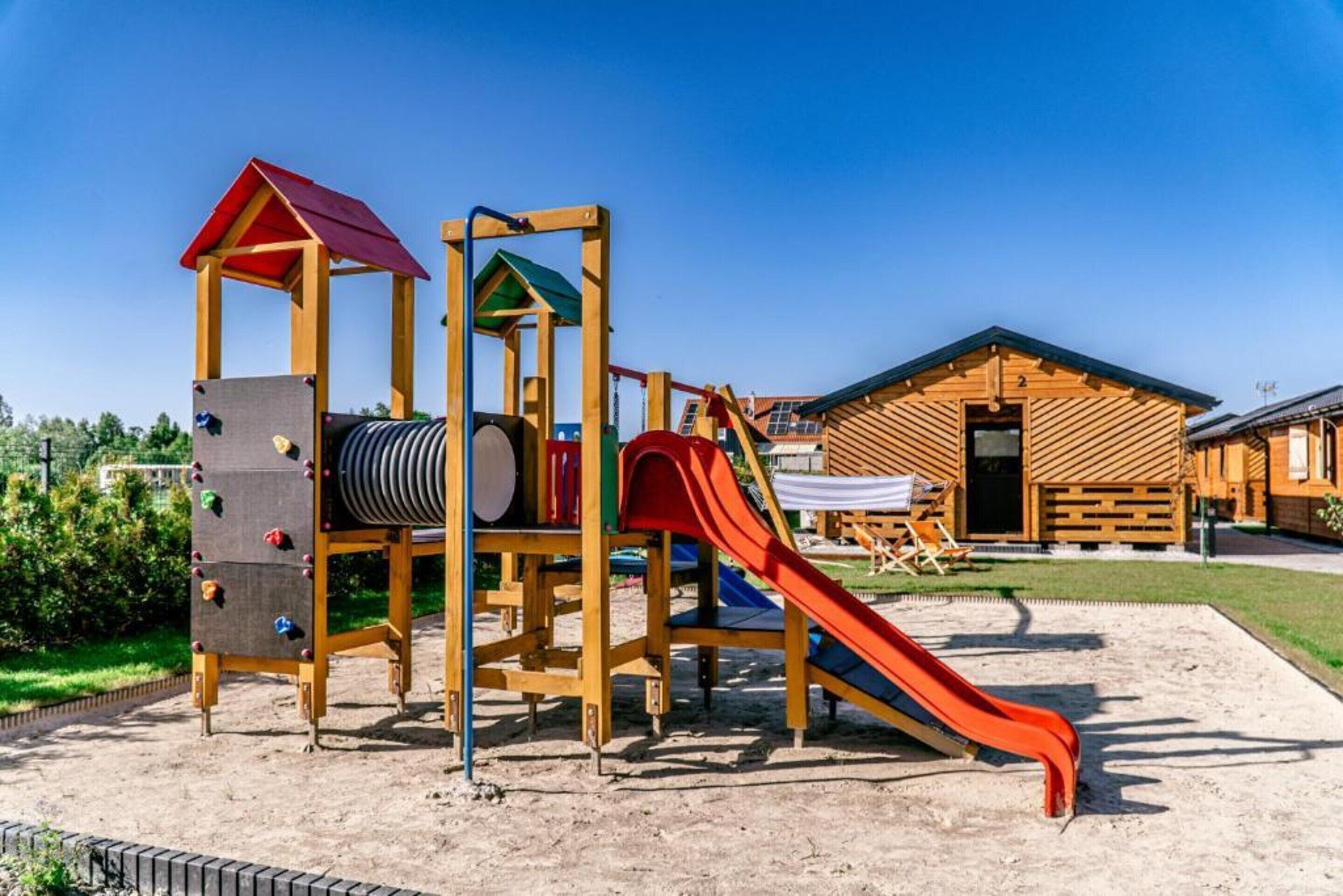 Un terrain de jeux avec des structures d’escalade et un toboggan, des cabanes en bois en arrière-plan et un ciel dégagé.