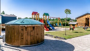 A wooden hot tub in a park with playground equipment in the background. - Holiday Homes With Terraces, Sauna + Whirlpool (gmina Będzino)