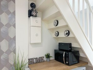 Eine kleine Küche mit Mikrowelle, einem Kühlschrank und einer Uhr an der Wand. Auf der Arbeitsfläche befinden sich ein Topf mit einer Pflanze und ein Willkommensschild.