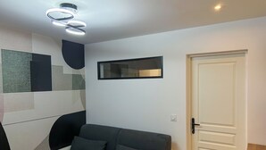 A modern living room with a dark grey sofa, a white door, and a geometric wall design. - Room N3 Colocation Confort & Service (91) (Saint-Pierre-du-Perray)