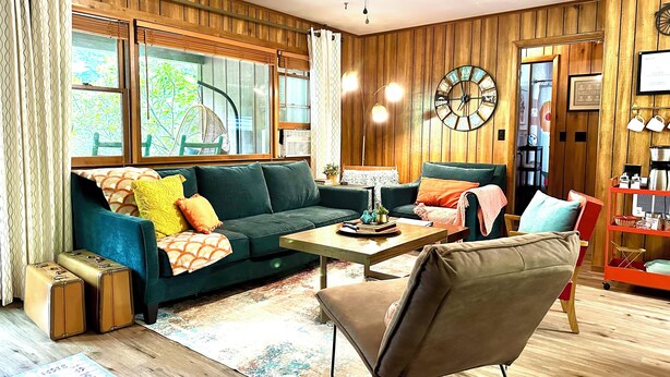 A cozy living room with a green sofa, a brown armchair, a wooden coffee table, and a large clock on the wall. - Smoky Mountain Creekside Retreat - Private Cabin with Backyard Stream (Maggie Valley)