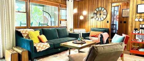 Una sala de estar acogedora con un sofá verde, un sillón marrón, una mesa de centro de madera y un reloj grande en la pared.