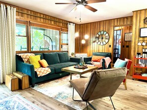 A cozy living room with a green sofa, a brown armchair, a wooden coffee table, and a large clock on the wall. - Smoky Mountain Creekside Retreat - Private Cabin with Backyard Stream (Maggie Valley)