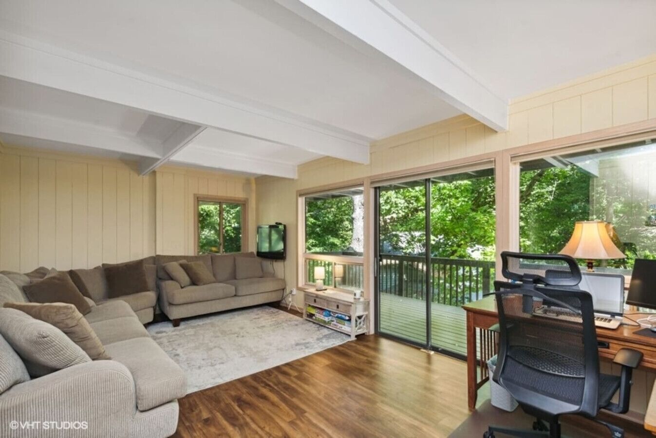 A living room with a sectional sofa, a desk with a computer, and a balcony with a view of trees.