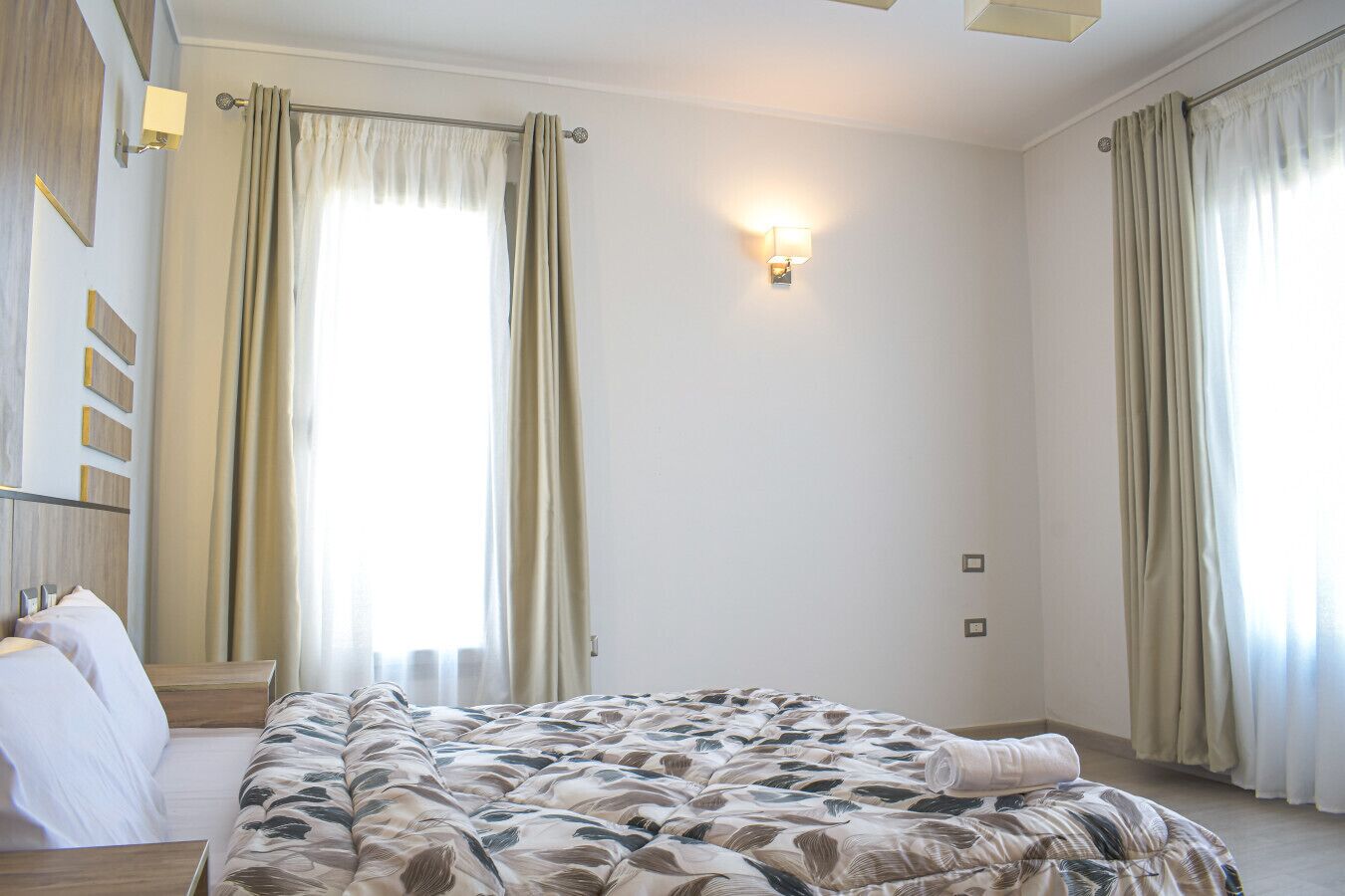 Ein Hotelzimmer mit einem großen Bett, zwei Nachttischen, einer Wandleuchte und einem fensterbepflanzten Fenster mit Vorhängen.