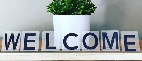 Una señal de bienvenida con letras en un estante, y una planta en maceta al fondo.