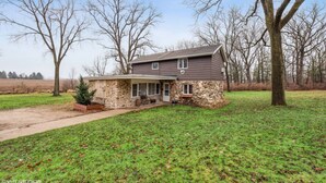 A two-story house with a stone foundation and a grey roof, surrounded by a grassy yard and leafless trees.