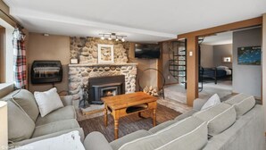 A cozy living room with a stone fireplace, a sofa set, a wooden coffee table, and a TV mounted on the wall.
