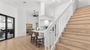 Eine moderne Küche mit weißen Schränken, einer zentralen Kochinsel mit Barhocker und einer Treppe mit Holzbeschlägen.