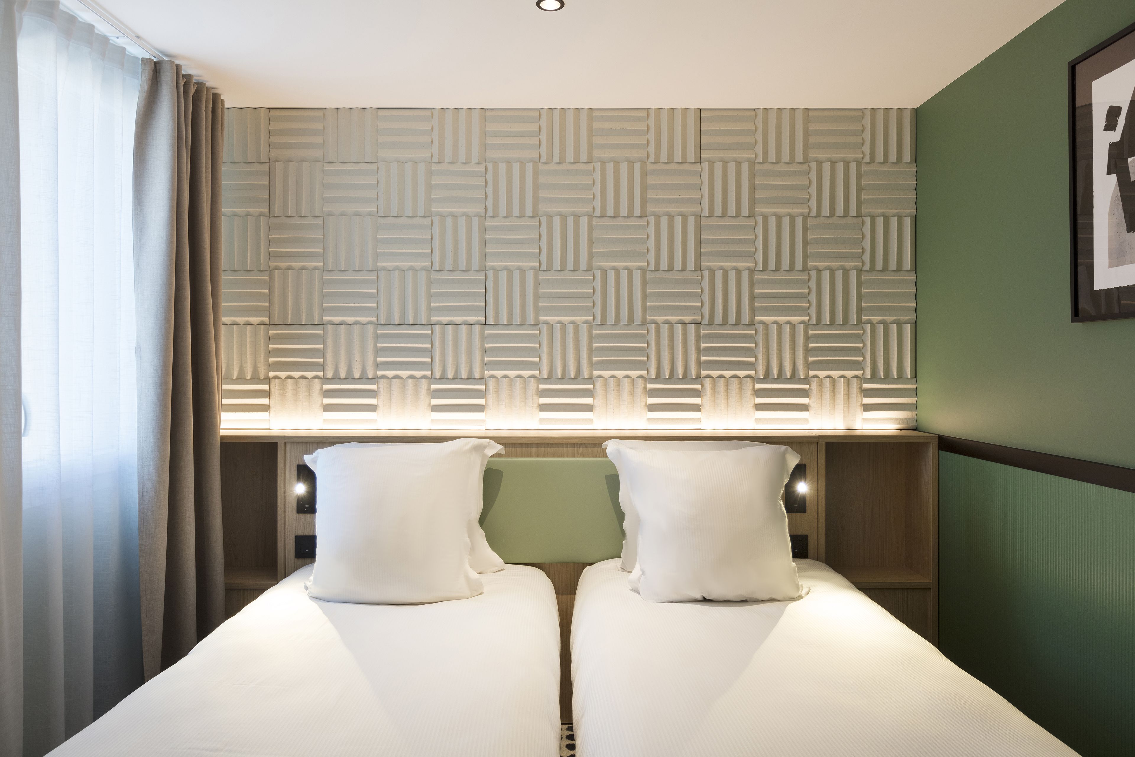 Een hotelkamer met een bed, een raam met gordijnen, een wand met een geometrisch patroon, en een ingelijst kunstwerk aan de wand.
