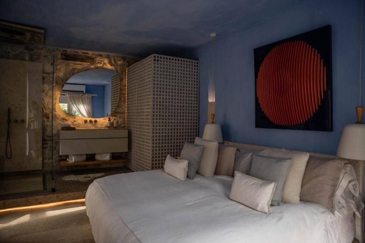 Une chambre avec un grand lit, un mur en pierre, une œuvre d’art moderne et une vue sur l’extérieur, visible à travers une arche.