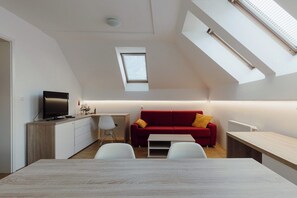 A modern loft apartment with a sloped ceiling, skylights, a red sofa, a dining table, and a TV. - Bright apartment with balcony and mountain view (Bled)