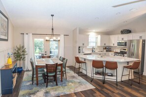 A modern kitchen with a central island, bar stools, and a dining area with a table and chairs. - Heated Pool, Golf Cart, Life Stlye ID's (The Villages)