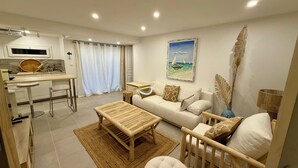 Ein modernes Wohnzimmer mit Sofa, Couchtisch und Babybett. Es gibt einen Essbereich mit Tisch und Stühlen sowie eine Küche mit Fenster.