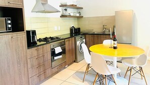 A modern kitchen with wooden cabinets, a microwave, a stove, a washing machine, and a yellow dining table with white chairs. - WELCOME TO 144 @ MAYFAIR — YOUR URBAN OASIS IN CENTURY CITY, CAPE TOWN! (Cape Town)