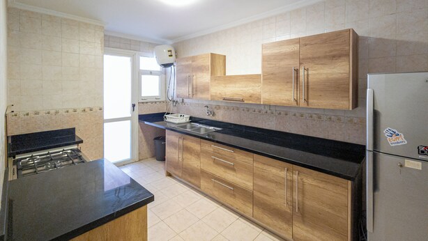 A kitchen with wooden cabinets, a black countertop, a stainless steel refrigerator, and a gas stove. - Pharaohs Residence New Cairo El Rehab (New Cairo El rehab)