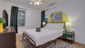 A bedroom with a bed, bedside tables, a ceiling fan, an air conditioner, a window with curtains, and a wall-mounted artwork. - 2BR Houston Haven, Playground, Prime Location (Houston)