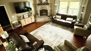 A living room with a fireplace, leather sofas, a flat-screen TV, and a large window with curtains. - Beautiful 4 bedroom Country Estate in Liberty. Minutes from downtown KC. (Liberty)