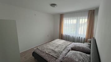 A small, neatly arranged bedroom with a single bed, a bedside table, and a window with sheer curtains.