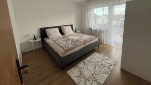 A bedroom with a bed, a bedside table, a nightstand, a window with sheer curtains, and a view of the outside.