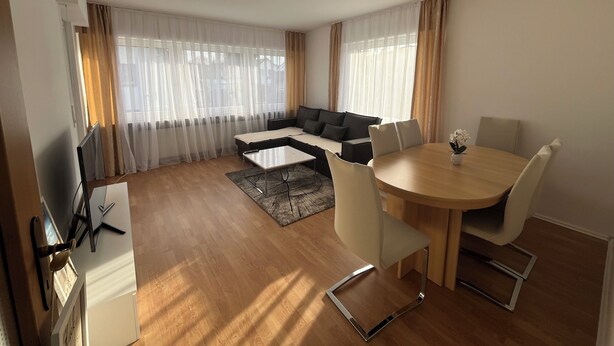 A modern living room with a sectional sofa, a dining table with chairs, and a flat-screen TV.