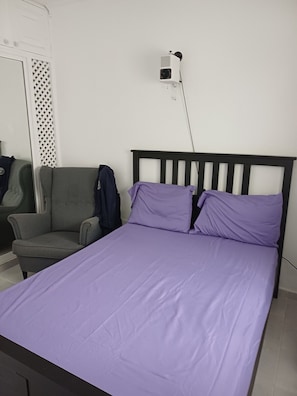 Ein Zimmer mit einem Einzelbett, einem Holz-Kopfteil, einem grauen Sessel und einer Wandsteckdose.
