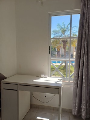 Ein Zimmer mit Schreibtisch, einem Fenster mit Blick auf Palmen und einen Pool, sowie einem Vorhang.