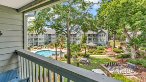 A balcony with a view of a swimming pool, a chessboard, and a well-maintained garden.
