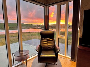 A room with a leather chair facing a large window overlooking a landscape with a body of water and fields.