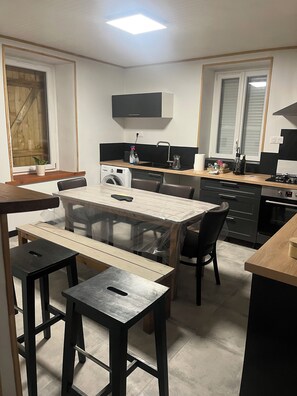 A compact kitchen with a dining area, a washing machine, and a window with wooden shutters. - Small fisherman's cottage (Loctudy)
