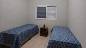 A small, clean room with two single beds, a window with blinds, and a small table with a plant. - CASA FLORENÇA (Indaiatuba)