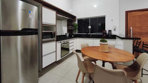 A modern kitchen with white cabinets, a stainless steel refrigerator, a microwave, and a stove. There is a dining table with chairs and a potted plant on it. - CASA FLORENÇA (Indaiatuba)