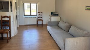 Une pièce avec un plancher en bois, un canapé gris, une chaise en bois, un radiateur, une fenêtre et un petit meuble blanc.