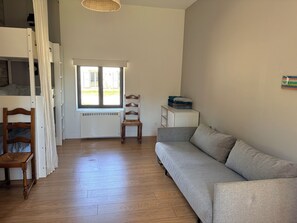Huoneessa on puulattia, harmaa sohva, puinen tuoli, patteri, ikkuna ja pieni valkoinen kaappi.