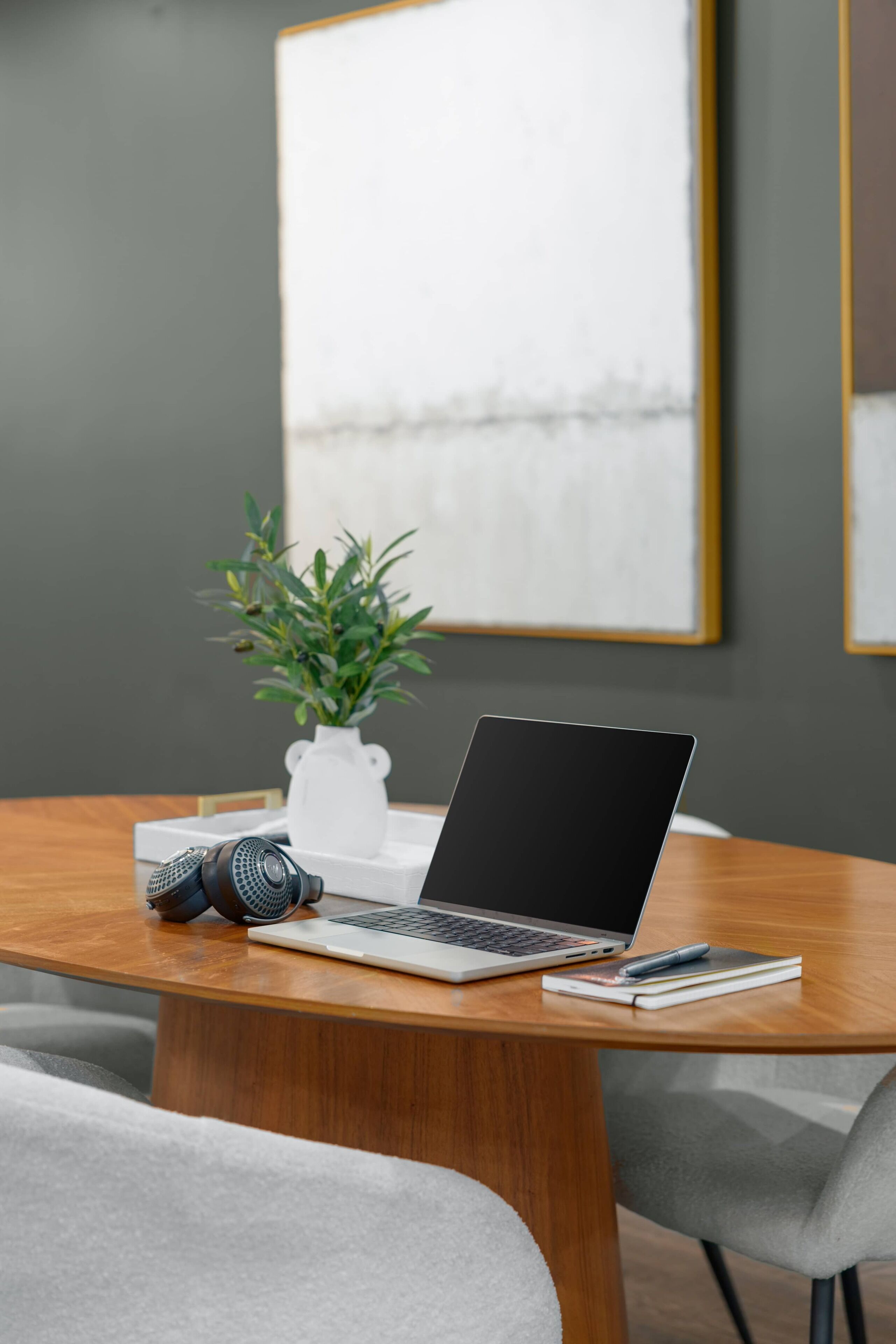 A laptop on a wooden table with a notebook, pen, and headphones beside it. A potted plant is on the table, and a framed picture hangs on the wall.