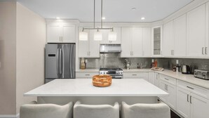 Une cuisine moderne avec des meubles blancs, un réfrigérateur en acier inoxydable, un îlot central doté de chaises et des luminaires suspendus.
