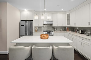 Una cocina moderna con armarios blancos, un refrigerador de acero inoxidable, una isla central con sillas e iluminación colgante.