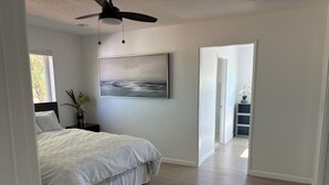 Une chambre à coucher avec un lit, un ventilateur de plafond, un tableau, une table de chevet et une fenêtre.