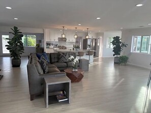 A spacious living room with a grey sectional sofa, a coffee table, and a bookshelf. There is a kitchen area with white cabinets and pendant lighting. - Spectacular lakeview, spacious, remodeled 3+2 bedroom, large lot for RVs, boats. (Perris)