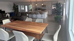 Un vaste salon avec une table à manger en bois, des chaises blanches, un canapé et une cuisine en arrière-plan.