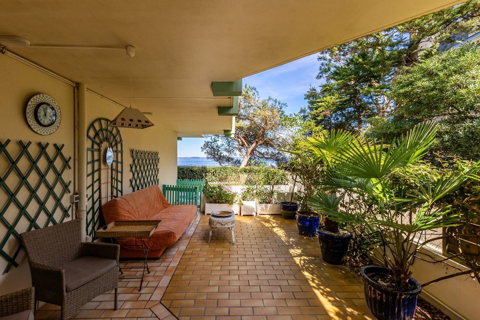 Ein überdachter Patio-Bereich mit einem Rattan-Sofa, einem kleinen Tisch und Topfpflanzen.