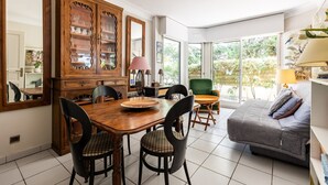 A dining area with a wooden table and chairs, a cabinet with glass doors, and a sofa in a bright room with large windows. - Emerald apartment with beach access in Pereire (Arcachon)
