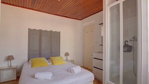 Een slaapkamer met een houten plafond, een bed met wit en geel beddengoed, twee nachtkastjes met lampen, een kledingkast, en een douchecel met glazen wanden.