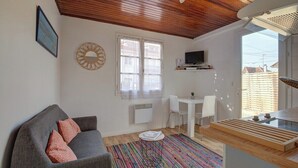 Een knus woongedeelte met een grijze bank, een klein rond tafeltje, een witte stoel, een kleurrijk gestreept vloerkleed, een houten plafond, een witte muur, een raam met witte gordijnen, een wandgemonteerde televisie en een klein plankje met een plant.