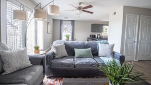 Una sala moderna con un sofá gris, dos sillones, un ventilador de techo y una cocina al fondo.
