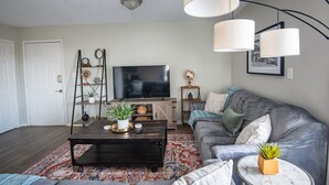 Una sala de estar con una mesa de centro de madera oscura, un sofá gris y un televisor sobre un mueble. Hay objetos decorativos en estanterías y una lámpara colgando del techo.