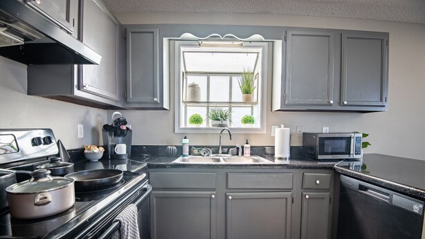 A kitchen with grey cabinets, a black countertop, a stove, a sink, a microwave, and a window with plants. - Ridge Retreats Unit B - Balcony/Wifi/4 Queen Beds (Birmingham)